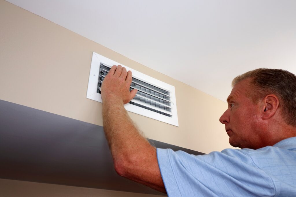 White rectangle air vent on a beige wall with a males hand on left side of the outflow furnace register. Mature male with left hand on and inspecting white home furnace air duct vent in a modern home
