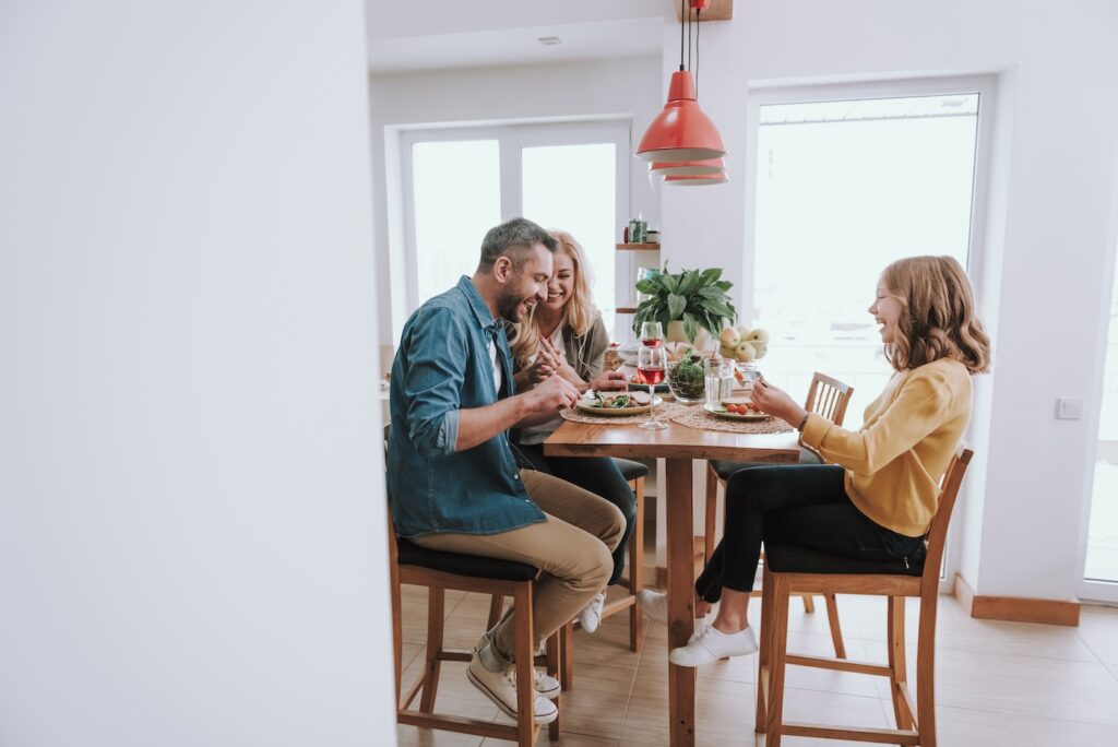 Side view portrait of joyful bearded man sitting at the kitchen table with his smiling wife and adorable daughter. Copy space in left side