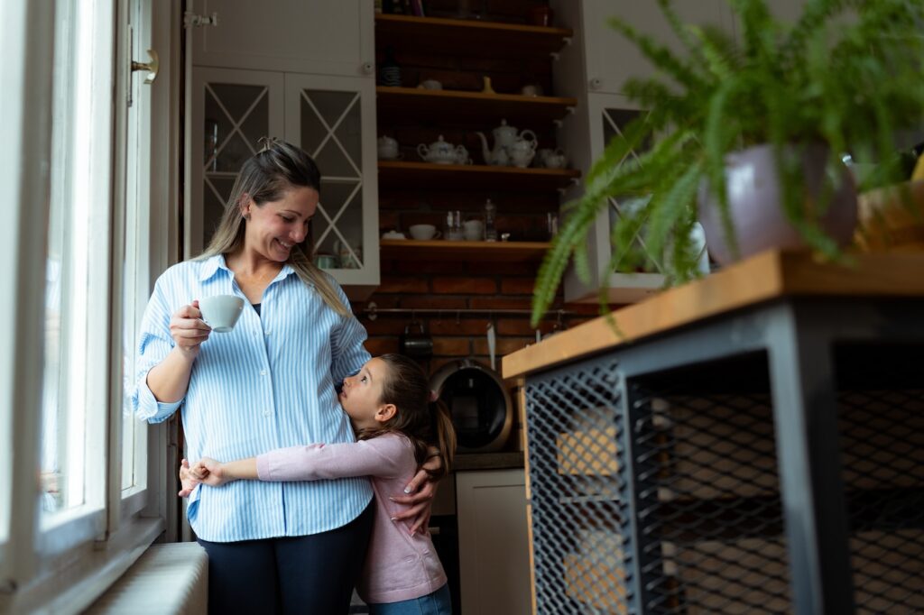 Mother enjoys a cup of coffee while her daughter hugs her affectionately in their modern kitchen, creating a heartwarming scene of family connection
