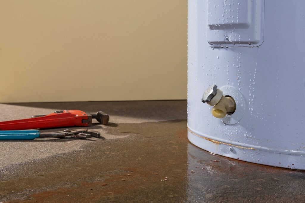 Water leaking from a residential electric water heater with a couple plumber’s tools sitting on the concrete floor.