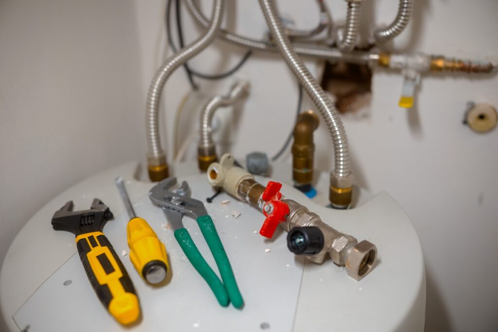 An adjustable wrench, screwdriver, and pliers rest on a white water heater amidst a network of metal pipes and valves
