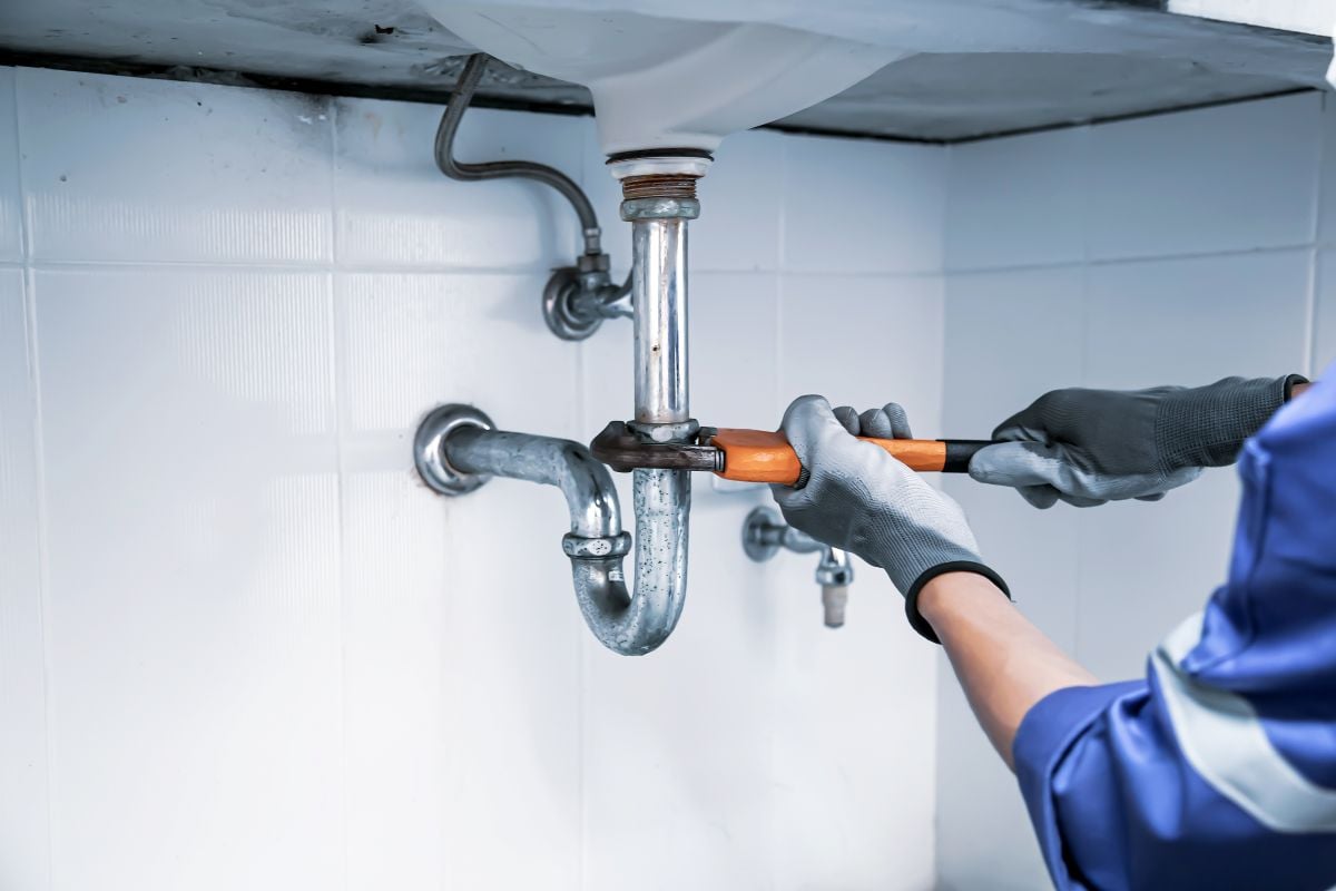 Plumbing worker with blue clothes and grey gloves fixing water pipe using wrench
